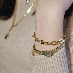 K&ouml;nnte beinhalten: Ein goldenes Kettenarmband mit geflochtenem Design und einem gro&szlig;en ovalen Stein. Das Armband ist &uuml;ber ein dickeres goldenes Kettenarmband gelegt.
