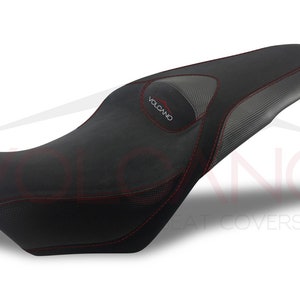 May include: Black motorcycle seat with red stitching and a carbon fiber pattern. The seat has the word "VOLCANO" in red. The seat is designed for a motorcycle and is likely an aftermarket part.