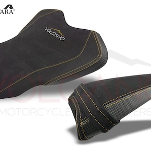 May include: A black motorcycle seat with yellow stitching and the word "VOLCANO" embroidered on it. The seat is made of Alcantara fabric and has a carbon fiber insert.