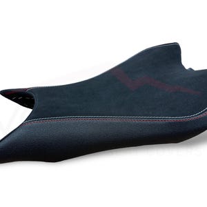 Puede incluir: Una funda de asiento de motocicleta negra con una superficie texturizada y costuras rojas. La funda del asiento tiene un panel central de gamuza con un dise&ntilde;o rojo. Las costuras blancas delinean los bordes.