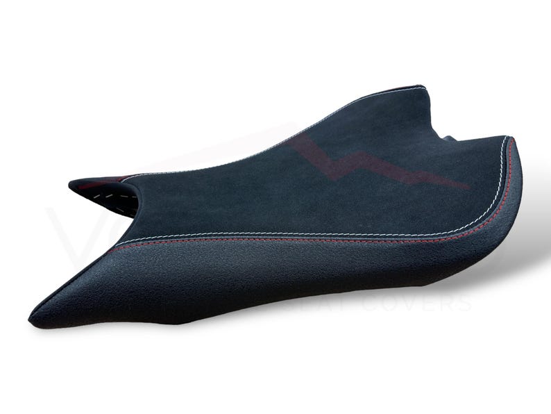 Puede incluir: Una funda de asiento de motocicleta negra con una superficie texturizada y costuras rojas. La funda del asiento tiene un panel central de gamuza con un dise&ntilde;o rojo. Las costuras blancas delinean los bordes.