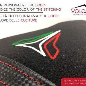 May include: Black motorcycle seat cover with red stitching. The seat cover features a white and green embroidered logo with the text "VOLCANO MOTORCYCLE SEAT COVERS". The text "YOU CAN PERSONALIZE THE LOGO AND CHOICE THE COLOR OF THE STITCHING POSSIBILIT&Agrave; DI PERSONALIZZARE IL LOGO E IL COLORE DELLE CUCITURE" is also visible.