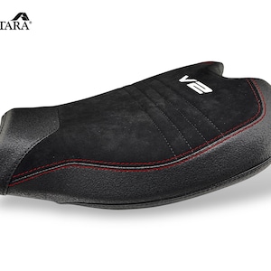 May include: Black motorcycle seat with red stitching and the text "V2" in white. The seat has a textured surface and is made of Alcantara material. The seat is designed for comfort and style.