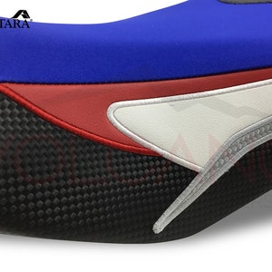 May include: A motorcycle seat cover featuring a blue, red and white design. The cover has a carbon fibre look and feel. The seat cover is made from Alcantara, a high-quality material.