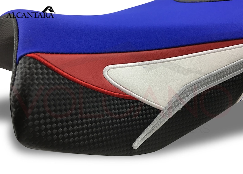 May include: A motorcycle seat cover featuring a blue, red and white design. The cover has a carbon fibre look and feel. The seat cover is made from Alcantara, a high-quality material.