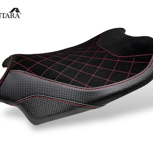 May include: Black motorcycle seat with a carbon fiber base and a diamond-stitched suede top. Red stitching accents the design. The brand name "ALCANTARA" is visible in the upper left corner.