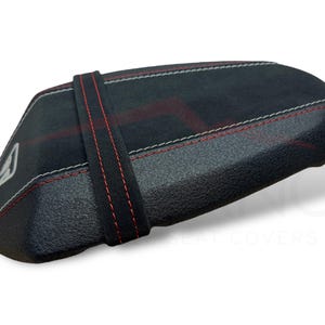 Puede incluir: Funda de asiento negra con costuras rojas y blancas. La funda tiene una superficie texturizada y una correa negra con costuras rojas. Un logotipo blanco es visible en el lateral.