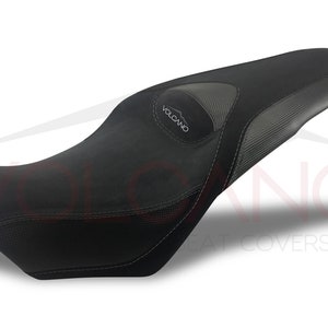 May include: Black motorcycle seat cover with a gray textured panel and the word "VOLCANO" in white lettering.