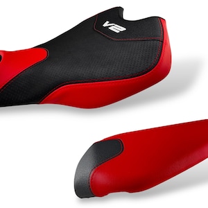May include: Two motorcycle seats, one with a black perforated top and red sides, the other all red with a black accent. The top seat has the text "V2" in white. Both seats are designed for a motorcycle.
