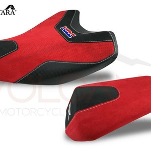 May include: A set of two motorcycle seat covers in red and black with a white stitching detail. The larger seat cover has the word "HRC" embroidered on it. The seat covers are made of Alcantara fabric.