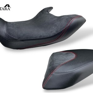 May include: Two black motorcycle seats with red stitching. The seats have a textured, suede-like center panel and a smooth, leather-like border. The brand name "ALCANTARA" is visible in the upper left corner.