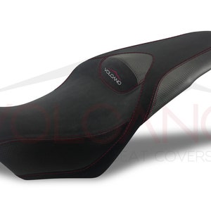 May include: Black motorcycle seat cover with red stitching and a gray carbon fiber panel. The seat cover has the word "VOLCANO" embroidered on it.