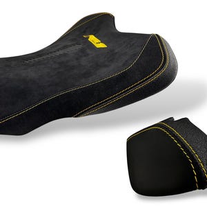 May include: Black motorcycle seat with yellow stitching and a yellow logo. The seat has a textured surface and a sleek design. A smaller matching piece is also shown.
