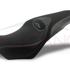 May include: Black motorcycle seat with a gray carbon fiber texture and white stitching. The seat has a raised section with the word "VOLCANO" in white text.