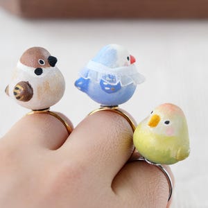 Könnte beinhalten: Drei goldene Ringe mit Vogelfiguren. Die Vögel sind ein brauner Spatz, ein blauer Vogel mit einem weißen Spitzenkragen und ein grün-gelber Vogel.