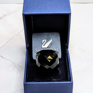 May include: A faceted black crystal ring displayed inside a dark blue hinged box. The interior of the box features a black velvet lining and a square plaque with the silver Swarovski swan logo and the text SWAROVSKI printed in white lettering.