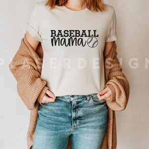 Op de afbeelding: Een wit T-shirt met de tekst "Baseball Mama" en een baseball-afbeelding gedrukt op de voorkant. Het shirt wordt gedragen door een persoon met een bruine cardigan en een blauwe jeans.