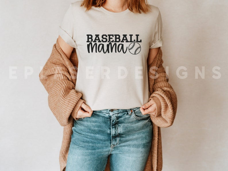 Op de afbeelding: Een wit T-shirt met de tekst "Baseball Mama" en een baseball-afbeelding gedrukt op de voorkant. Het shirt wordt gedragen door een persoon met een bruine cardigan en een blauwe jeans.