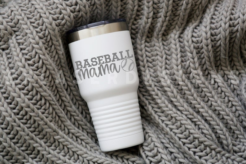 Op de afbeelding: Een witte roestvrijstalen beker met een grijze baseball en de tekst "Baseball mama" erop. De beker staat op een grijs gebreid deken.
