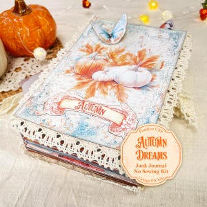 Op de afbeelding: Een junk journal kit met een omslag met een illustratie van pompoenen en herfstbladeren, een blauwe vlinderuitsnede en crèmekleurige kanten rand. De omslag toont de tekst "AUTUMN" en een label met "Positive Chic AUTUMN DREAMS Junk Journal No Sewing Kit".