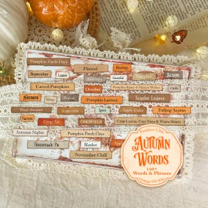 May include: A collection of rectangular paper labels featuring autumn-themed words and phrases such as Pumpkin Patch Days, Flannel, Burlap, October, and Crisp Leaves, Cozy Days & Warm Hearts, presented on a sheet with a decorative lace border.