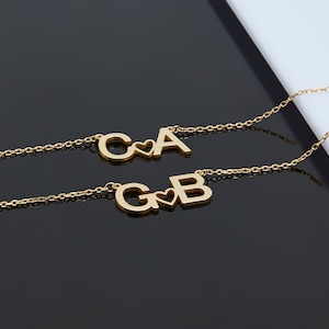 May include: Two gold necklaces with initial charms. The first necklace has the letters "C" and "A" with a heart between them. The second necklace has the letters "G" and "B" with a heart between them.