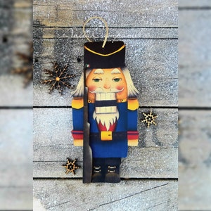 Puede incluir: Un adorno de cascanueces de madera pintado en azul, rojo y amarillo con barba y bigote blancos. El cascanueces sostiene un rifle y lleva un sombrero negro con ribete dorado.