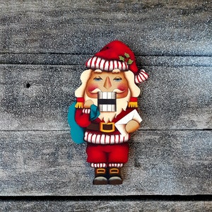Puede incluir: Un adorno de cascanueces de madera pintado para parecerse a Santa Claus. Lleva un gorro rojo y blanco, un abrigo a rayas rojo y blanco y una bolsa azul. Lleva una carta en su mano derecha.