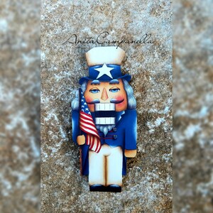 Puede incluir: Un cascanueces de madera pintado para parecerse al Tío Sam. Lleva una chaqueta azul, pantalones blancos y un sombrero rojo, blanco y azul. Sostiene una pequeña bandera estadounidense en su mano derecha. El cascanueces está de pie sobre una superficie marrón texturizada. Anita Campanella está escrito en cursiva en la parte superior de la imagen.