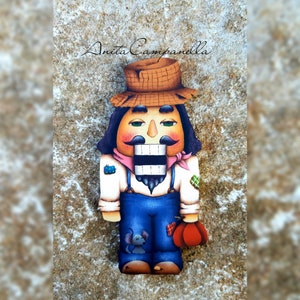 Puede incluir: Una figura de cascanueces de madera pintada a mano con un sombrero marrón, un mono azul y una bufanda rosa y blanca. El cascanueces tiene bigote y barba, y sostiene un bastón de caramelo rojo y blanco.