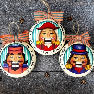 Puede incluir: Tres adornos redondos de madera blanca con caras de cascanueces pintadas y lemas navideños festivos. Los adornos están decorados con rayas azul verdoso y blancas y lazos de cinta naranja y blanca. Los lemas son "Dreaming with Faith in the Heart of Christmas", "The Magic of Christmas Nutcracker" y "Christmas Night is this time".
