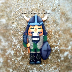 Puede incluir: Una figura de cascanueces de madera pintada en verde, azul y negro. El cascanueces lleva un casco con cuernos y sostiene un hacha y un escudo. El cascanueces tiene bigote y una sonrisa de dientes.