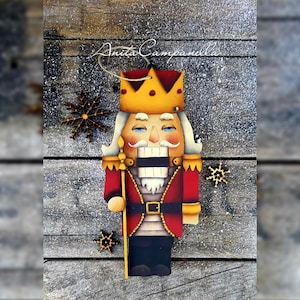 Puede incluir: Un adorno de cascanueces de madera pintado a mano con un uniforme rojo y dorado, una corona y un bigote. El adorno está decorado con copos de nieve y tiene un fondo de madera rústico.