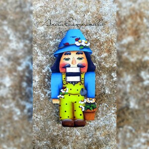 Puede incluir: Una figurita de cascanueces de madera pintada en un estilo caprichoso. El cascanueces lleva un sombrero azul con flores blancas, una camisa azul y un mono verde con lunares blancos. El cascanueces sostiene una pequeña maceta marrón con una planta verde.