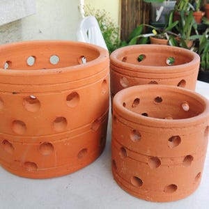 Könnte beinhalten: Drei unterschiedlich große Blumentöpfe aus Terrakotta. Jeder Topf ist zylindrisch mit einer Reihe von gleichmäßig verteilten Löchern an den Seiten. Die Töpfe haben eine warme, rötlich-braune Farbe und eignen sich für den Innen- und Außenbereich.