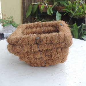 May include: A square coconut fibre planter, suitable for gardening. The planter is a warm brown colour and is constructed with layers of coconut fibre, held together with wire. It sits on a white surface, ready for planting.