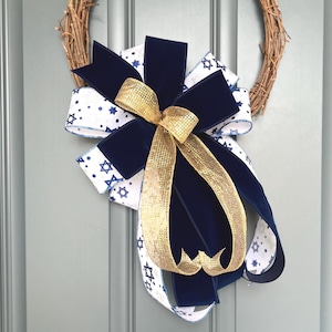 May include: A decorative bow featuring layers of navy blue velvet ribbon, white ribbon with blue Star of David and dot patterns, and a central gold metallic mesh ribbon. The bow is attached to a circular brown twig wreath base.