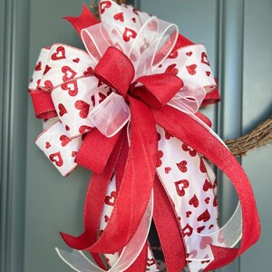 May include: A decorative bow made from layered ribbons featuring a white background with red glittery heart patterns and solid red fabric ribbons. The bow has multiple loops and long trailing tails, creating a full, multi-dimensional appearance.