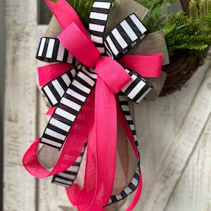 May include: A decorative wreath featuring a vibrant pink bow with black and white striped ribbon accents. The wreath includes a brown burlap ribbon and is adorned with green foliage, creating a cheerful and stylish home decor piece.