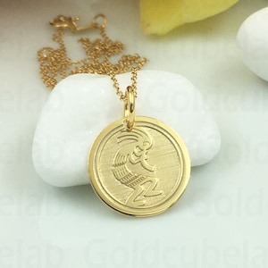 May include: A gold-colored pendant necklace featuring a circular charm with a Kokopelli design. The pendant hangs from a delicate gold chain. The necklace is displayed on a white stone, with flowers in the background.