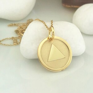 May include: A gold necklace with a round pendant featuring a gold triangle inside. The pendant is on a white surface.