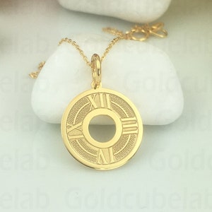 May include: A gold-colored pendant necklace featuring a circular pendant with Roman numerals and a central design. The pendant hangs from a delicate gold chain. The necklace is displayed on a white surface.