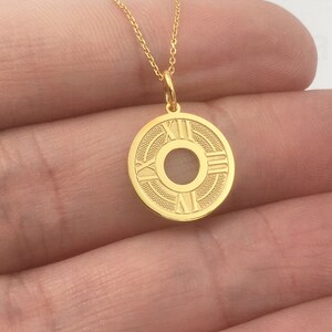 May include: A gold-colored pendant necklace featuring a circular charm with Roman numerals and a central design. The charm is suspended from a delicate gold chain. The necklace is displayed against a neutral background.