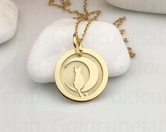 Peut inclure: Un collier pendentif doré avec un charme circulaire représentant un chat sur un croissant de lune. La chaîne est fine et le bijou est présenté sur des pierres blanches, avec des fleurs en arrière-plan. Le pendentif est en or.