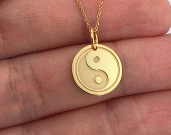 Puede incluir: Un collar dorado con un colgante circular con el símbolo del Yin y Yang. El colgante cuelga de una fina cadena dorada. El collar se muestra sobre un fondo claro.