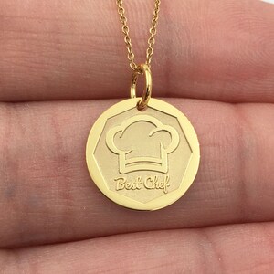 May include: A gold-colored necklace with a pendant featuring a chef's hat design and the words "Best Chef". The pendant is round with a geometric border and hangs from a delicate chain. The necklace is a culinary-themed accessory.