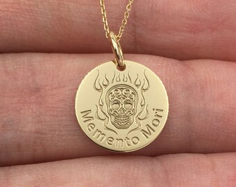 May include: A gold-colored pendant necklace featuring a circular charm with a skull design and the words "Memento Mori." The skull is surrounded by flames. The necklace is displayed on a light background.