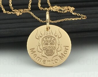 May include: Gold-colored pendant necklace featuring a circular pendant with a skull design and the words "Memento Mori". The skull is surrounded by flames. The necklace is displayed on a black surface.