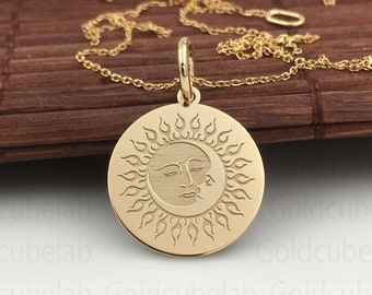 May include: Gold-colored pendant necklace featuring a circular pendant with a sun and moon design. The sun has a face and is surrounded by flames. The necklace chain is delicate and gold-toned, resting on a wooden surface.