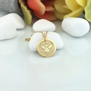 May include: A gold-colored pendant necklace featuring a circular charm with an angel design. The angel has outstretched wings and a halo. The necklace is displayed on a white surface with white stones and colorful flowers in the background.